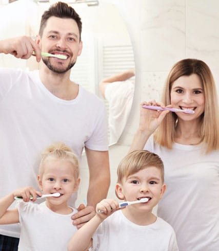 Family of four brushing teeth together in bathroom, smiling at camera.