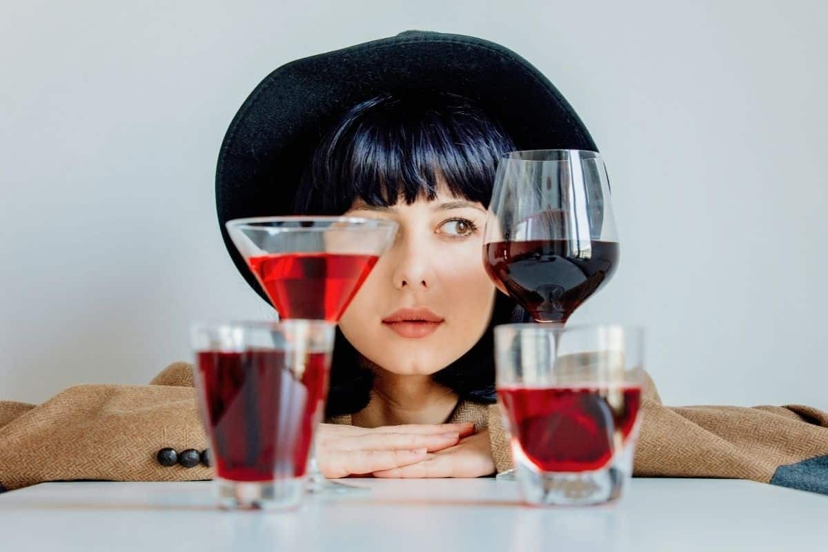 A woman wearing a hat looks past several glasses of red wine