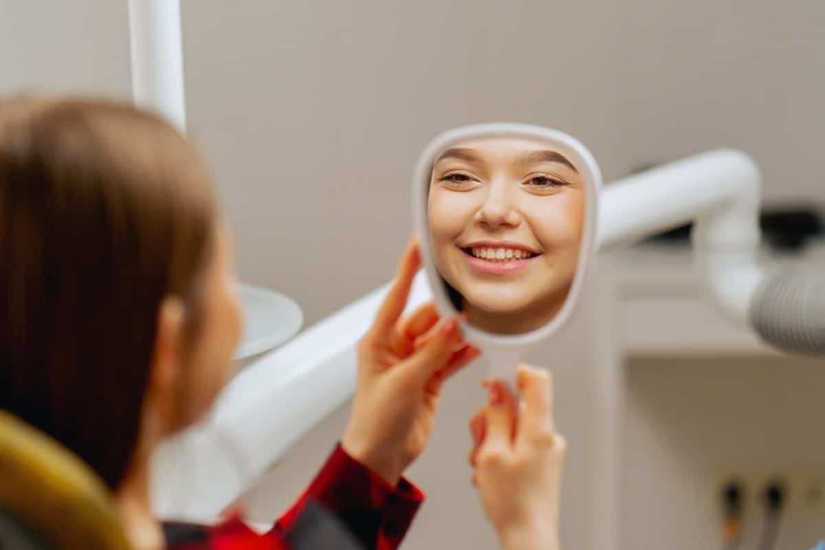 Smiling teen looking at her reflection in a handheld mirror at a dental clinic.